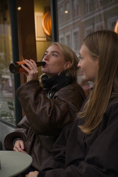 Two women sitting together, one drinking from a bottle of Good Orange outside a cafe