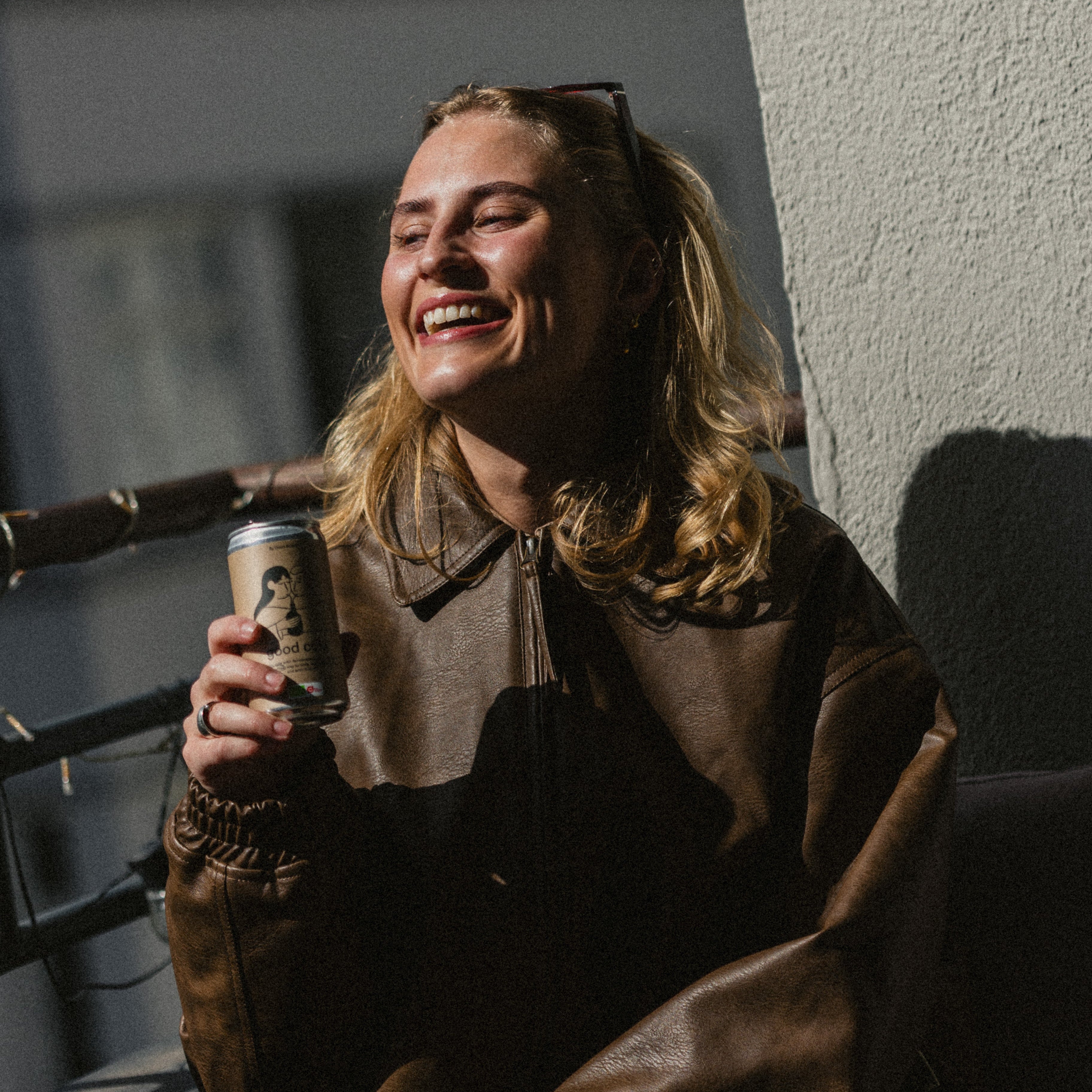 A woman sitting on a balcony holding a can of the fermentet and organic good cola, laughing 