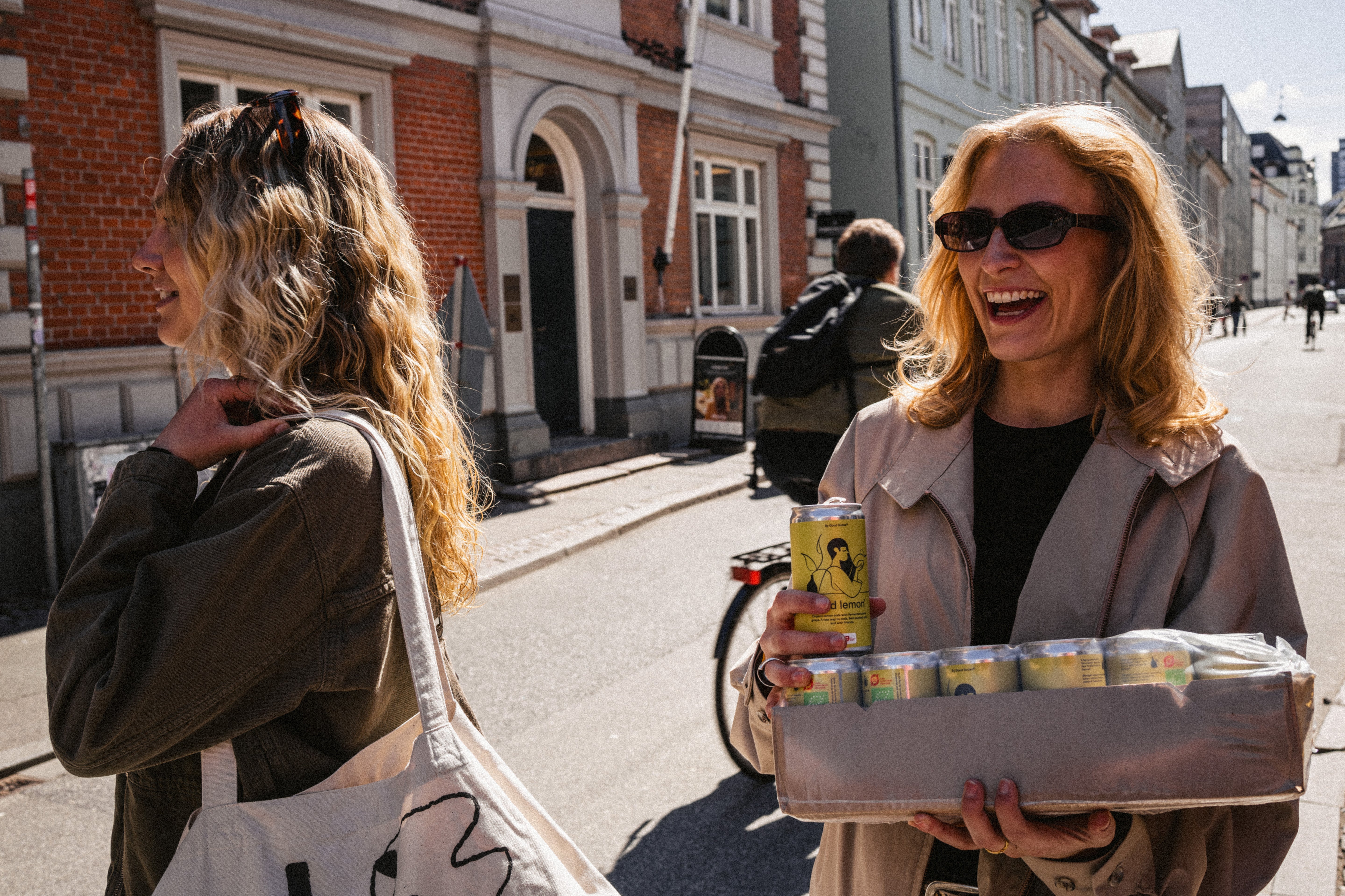 Two women on a street, one holding a box of our organic and fermentet good lemon® soda  and the other with a bag.