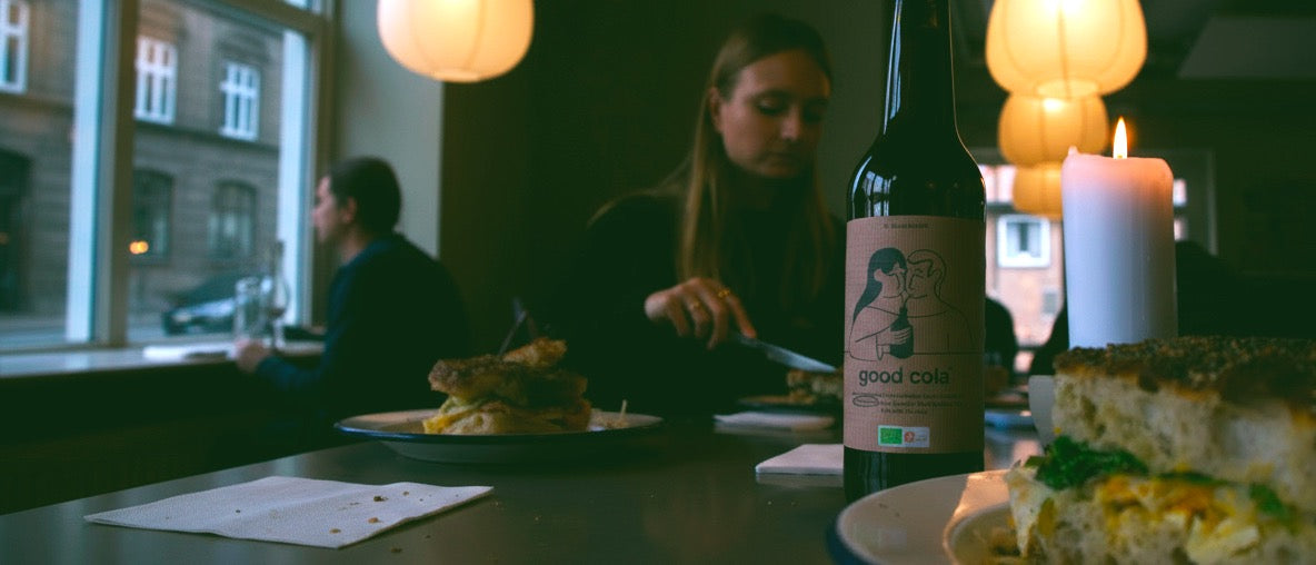 Person sitting at a table with a bottle  of organic and fermentet 'good cola' in a restaurant setting.