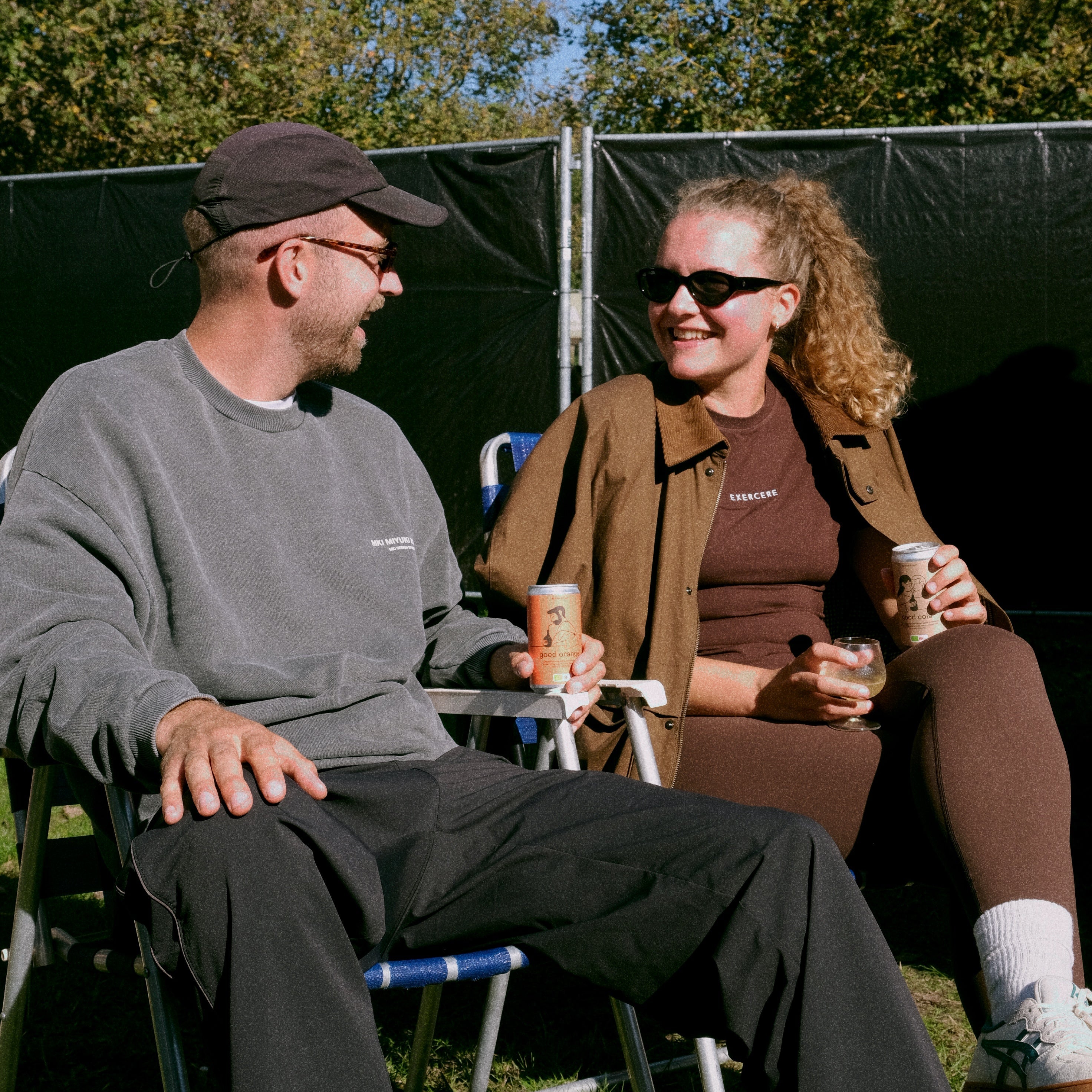 a man and a woman drinking the fermentet and organic good orange soda sitting in camping chairs outdoors at a festival