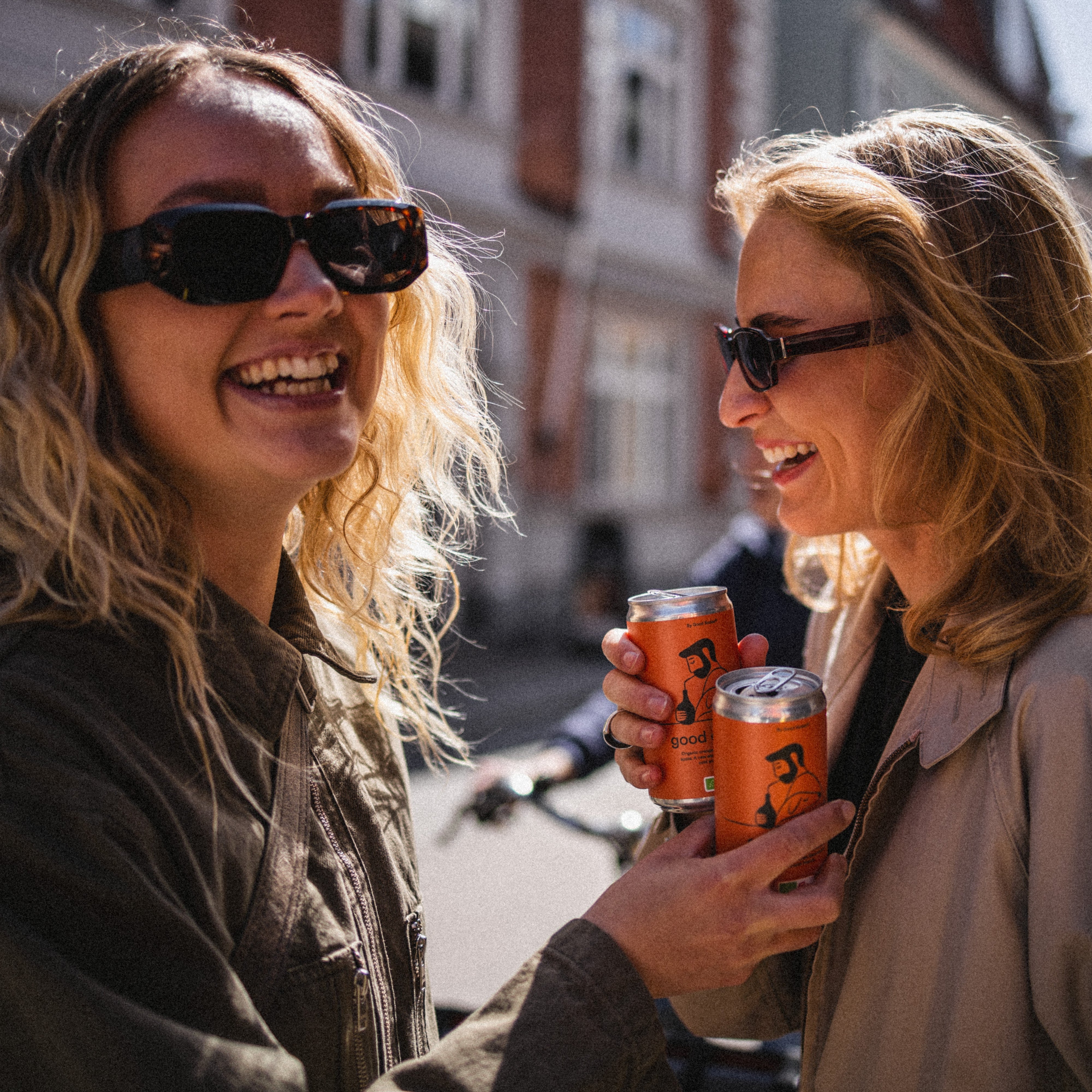 Two women standing on a street, holding cans of the organic and fermentet good orange soda and smiling.